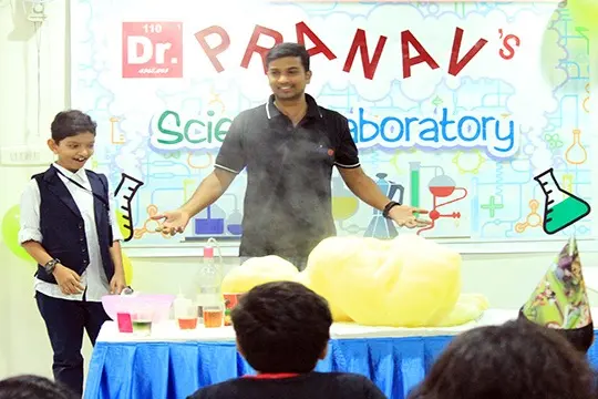 A host and a child at a decorated science table as a tall column of foam erupts