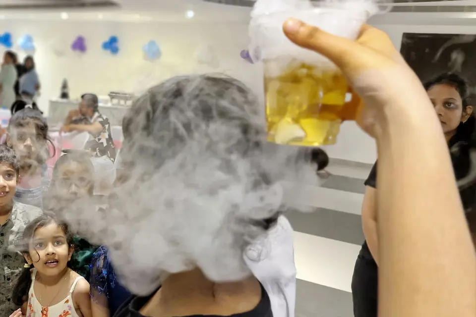 A hand tipping a jug of dry-ice fog as a row of children watch the smoke pour out