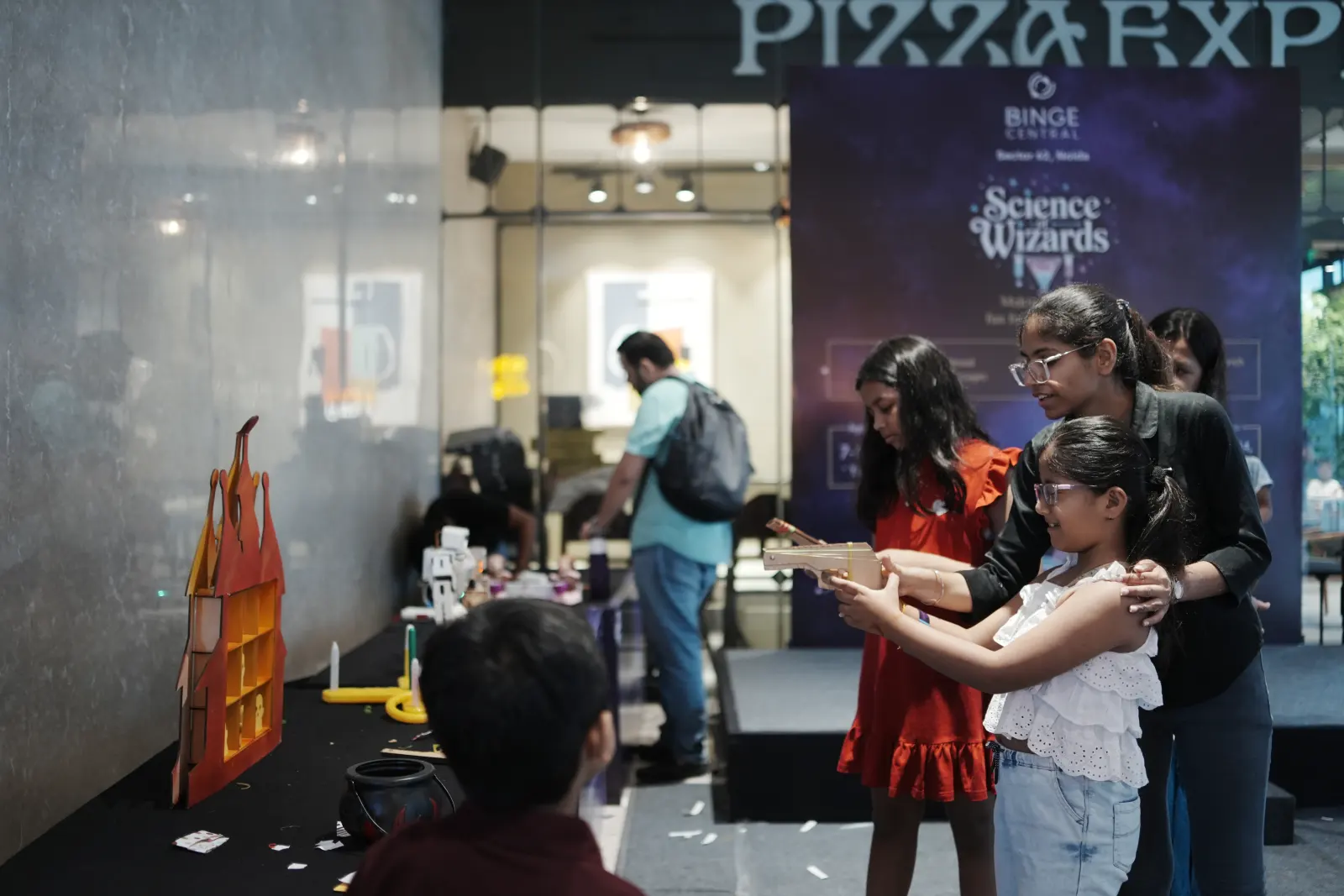 A mother steadying her daughter’s aim at a wooden castle target game during a Science Wizards event