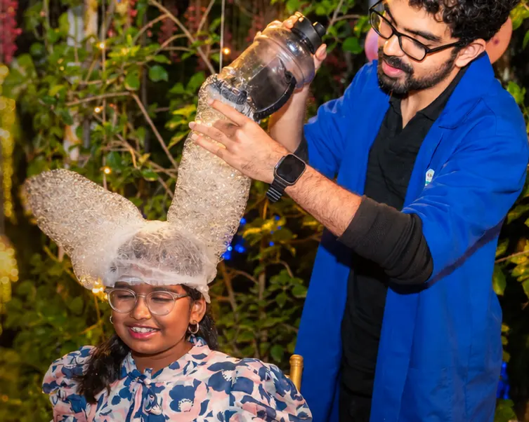 A MakerMasti host pouring a tower of bubble foam onto a laughing girl’s head at a garden birthday party