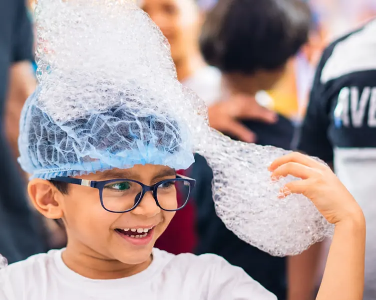A boy laughing with a tower of bubble foam on his head at a MakerMasti science activity