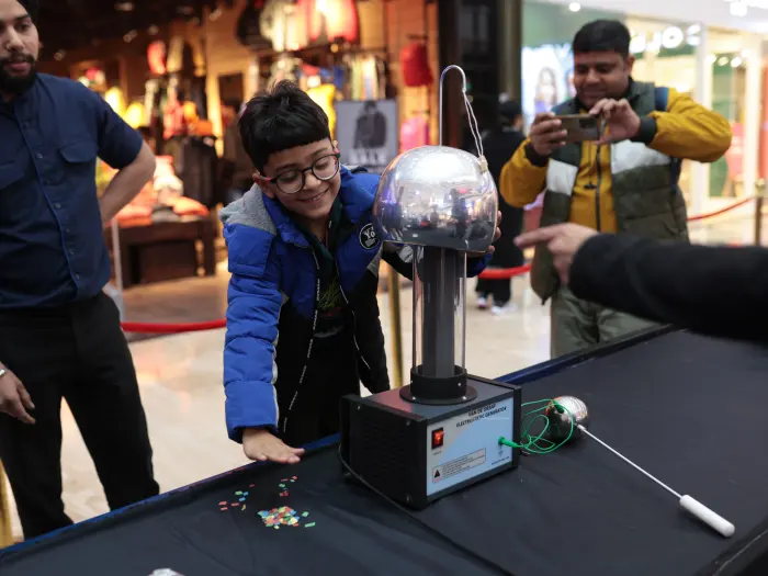 A boy grinning as he touches a Van de Graaff generator while shoppers stop to watch at a mall science festival